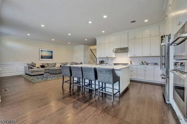 kitchen with decorative backsplash, appliances with stainless steel finishes, under cabinet range hood, white cabinetry, and a kitchen breakfast bar