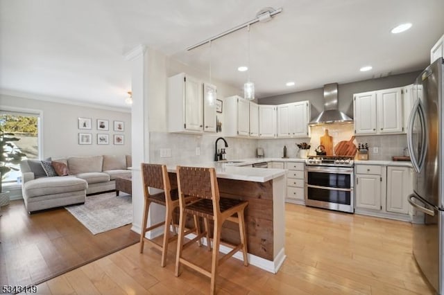 kitchen with a peninsula, a sink, stainless steel appliances, wall chimney range hood, and open floor plan