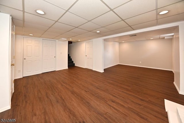 finished below grade area featuring a paneled ceiling, baseboards, dark wood finished floors, and stairs