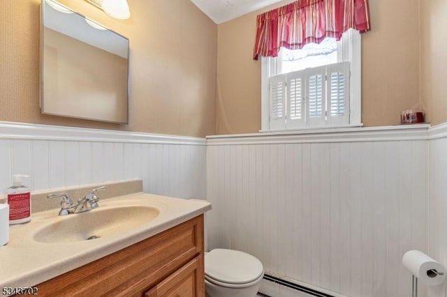 half bathroom with a baseboard heating unit, vanity, wainscoting, and toilet