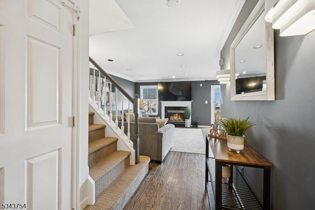 staircase featuring recessed lighting, a warm lit fireplace, crown molding, and hardwood / wood-style floors