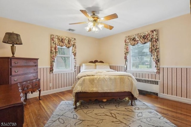 bedroom featuring visible vents, ceiling fan, radiator heating unit, wainscoting, and wood finished floors