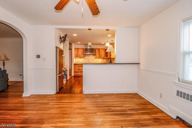 kitchen featuring wall chimney range hood, freestanding refrigerator, wood finished floors, arched walkways, and a ceiling fan