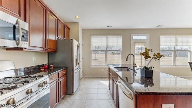 kitchen featuring an island with sink, a sink, dark stone countertops, stainless steel appliances, and light tile patterned floors