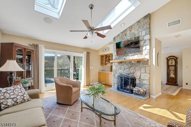 living room with light wood-type flooring, a ceiling fan, a stone fireplace, visible vents, and a skylight