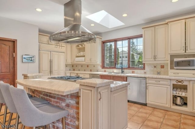 kitchen featuring tile counters, appliances with stainless steel finishes, a skylight, island range hood, and a sink