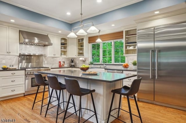 kitchen featuring high end appliances, wall chimney exhaust hood, dark countertops, ornamental molding, and a kitchen bar