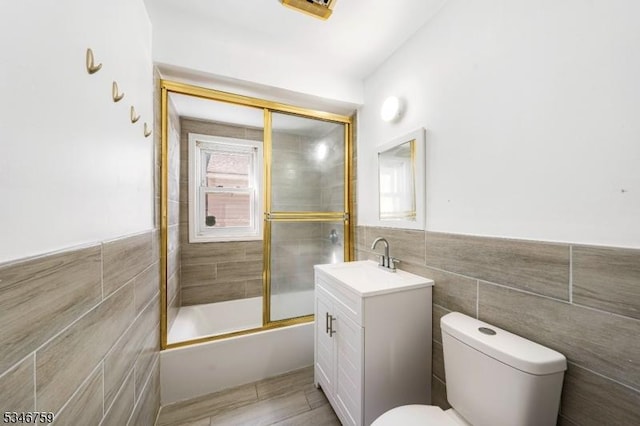 full bathroom with bath / shower combo with glass door, toilet, vanity, and tile walls