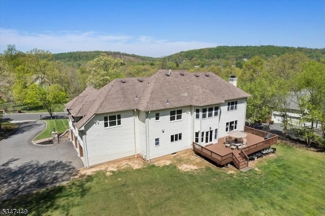 rear view of house featuring a wooden deck, roof with shingles, a chimney, a lawn, and a forest view