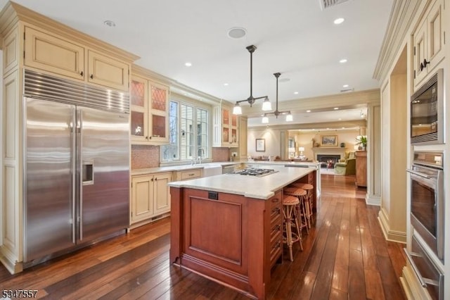 kitchen with glass insert cabinets, built in appliances, light countertops, cream cabinetry, and dark wood-style flooring