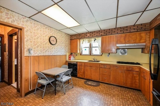 kitchen with wallpapered walls, black appliances, under cabinet range hood, a drop ceiling, and a wainscoted wall