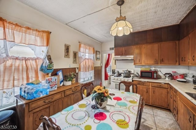 kitchen with light tile patterned floors, light countertops, under cabinet range hood, brown cabinetry, and white gas range oven