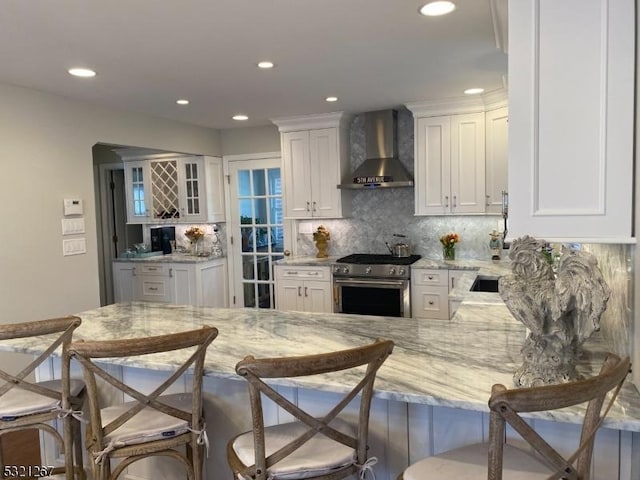 kitchen featuring wall chimney range hood, light stone countertops, stainless steel range, and white cabinetry