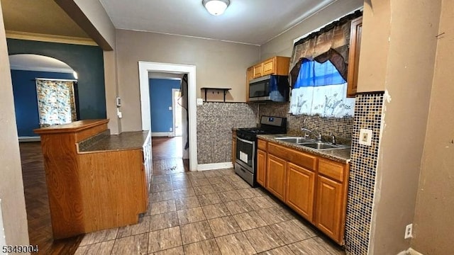 kitchen with brown cabinetry, dark countertops, appliances with stainless steel finishes, arched walkways, and a sink