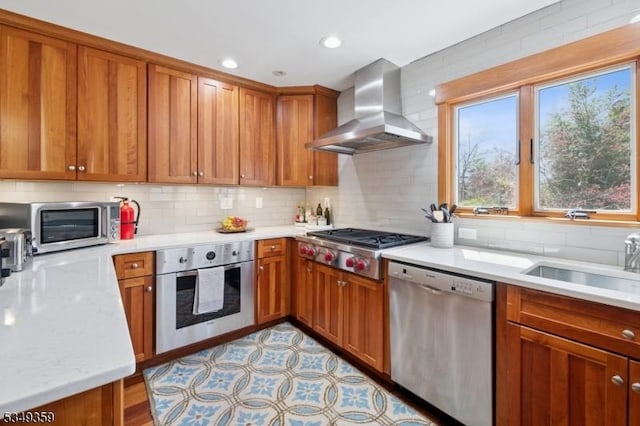 kitchen featuring wall chimney range hood, appliances with stainless steel finishes, a sink, brown cabinetry, and tasteful backsplash