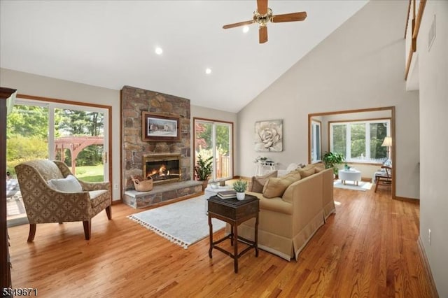 living area featuring high vaulted ceiling, light wood-type flooring, ceiling fan, a stone fireplace, and baseboards
