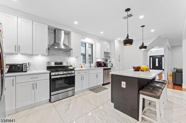 kitchen with a kitchen breakfast bar, wall chimney range hood, white cabinetry, a kitchen island, and appliances with stainless steel finishes
