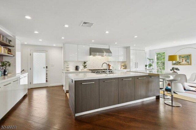 kitchen with modern cabinets, dark wood-style floors, wall chimney range hood, white cabinetry, and a large island with sink