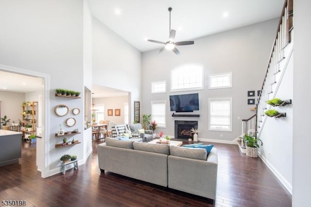 living area featuring ceiling fan, dark wood finished floors, a towering ceiling, stairs, and a glass covered fireplace