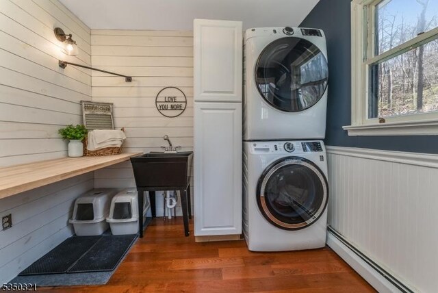 clothes washing area featuring a baseboard heating unit, wood walls, cabinet space, and stacked washer / drying machine