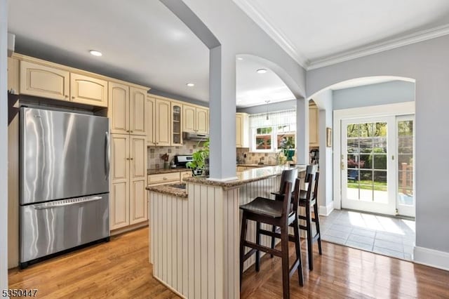 kitchen featuring crown molding, freestanding refrigerator, stone counters, cream cabinetry, and backsplash