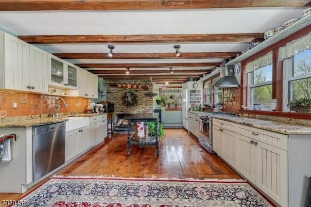 kitchen with light wood-type flooring, wall chimney exhaust hood, appliances with stainless steel finishes, and glass insert cabinets