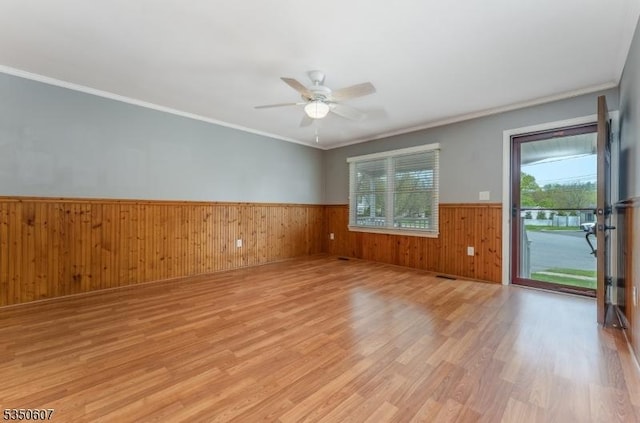 empty room with crown molding, wood walls, wainscoting, a ceiling fan, and light wood-style flooring