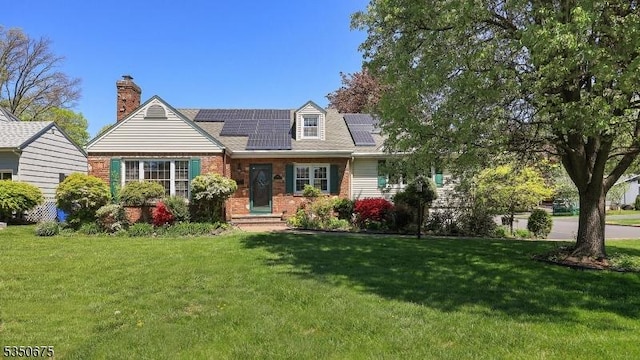 cape cod house with brick siding, a chimney, a front yard, and solar panels