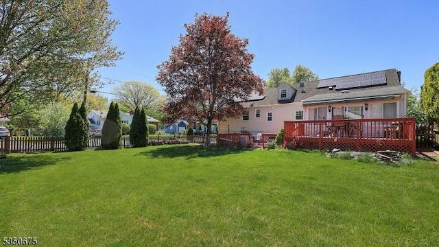 back of property featuring fence, a shingled roof, a wooden deck, roof mounted solar panels, and a lawn