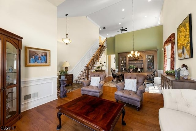 living area featuring high vaulted ceiling, ceiling fan with notable chandelier, a wainscoted wall, stairs, and wood finished floors