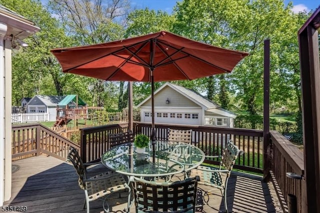 wooden terrace featuring outdoor dining area, an outbuilding, a detached garage, fence, and a playground