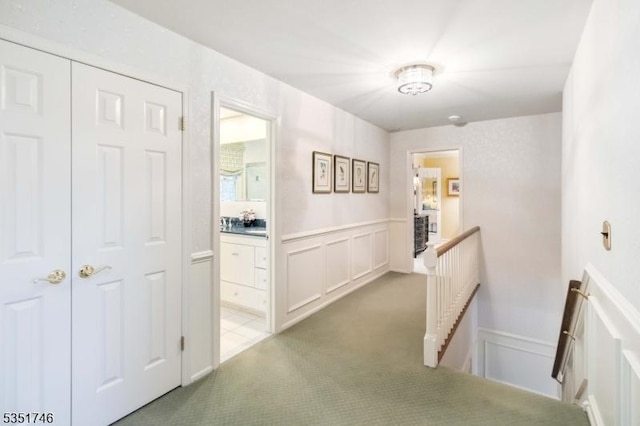 corridor with light colored carpet, a decorative wall, an upstairs landing, and a wainscoted wall