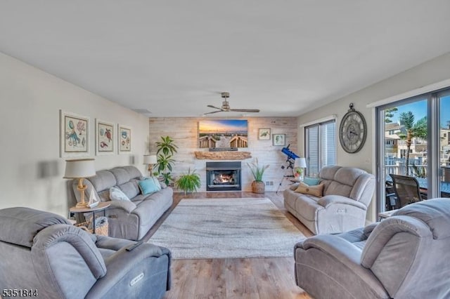 living room featuring wood walls, ceiling fan, a warm lit fireplace, and wood finished floors