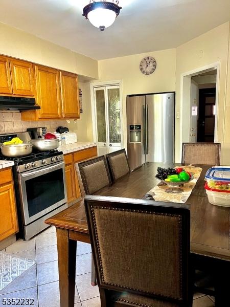 kitchen with light tile patterned flooring, decorative backsplash, brown cabinetry, under cabinet range hood, and appliances with stainless steel finishes