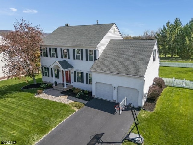 colonial-style house featuring a front lawn, aphalt driveway, fence, a garage, and roof with shingles