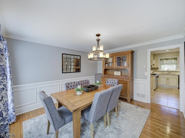 dining space featuring a chandelier, light wood-type flooring, ornamental molding, a wainscoted wall, and a decorative wall
