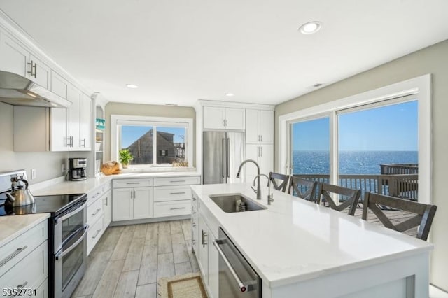kitchen featuring a center island with sink, light wood-type flooring, under cabinet range hood, a sink, and appliances with stainless steel finishes