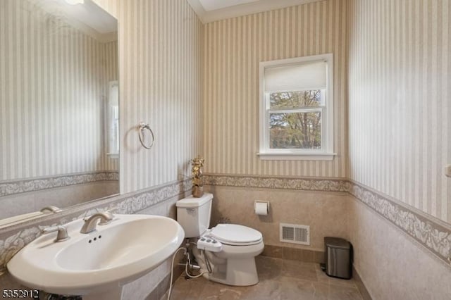 bathroom featuring a sink, toilet, wainscoting, visible vents, and wallpapered walls