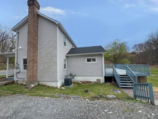 rear view of property with a wooden deck, a chimney, roof with shingles, a lawn, and central AC unit
