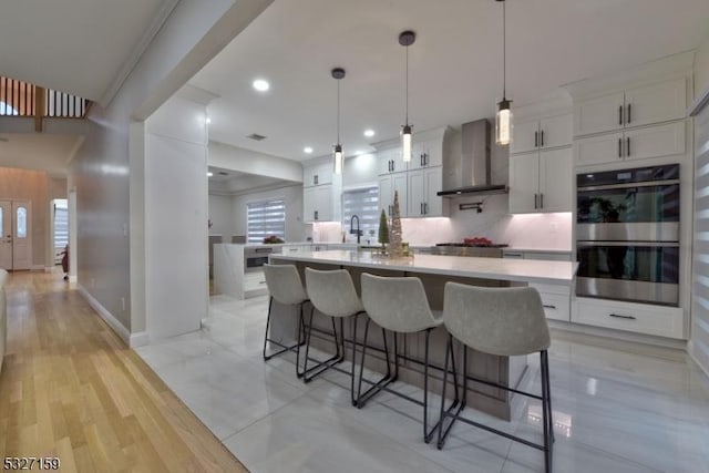 kitchen with double oven, white cabinetry, light countertops, and wall chimney exhaust hood