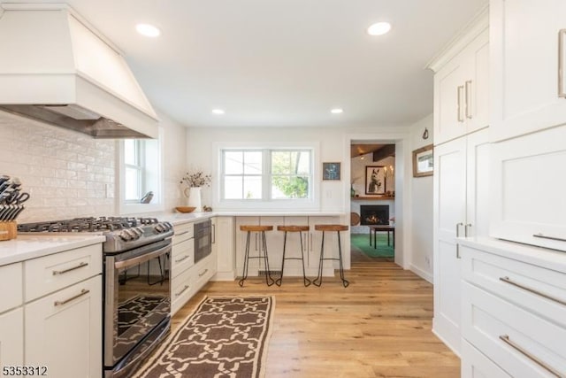 kitchen featuring stainless steel range with gas cooktop, custom exhaust hood, light wood-style flooring, backsplash, and light countertops