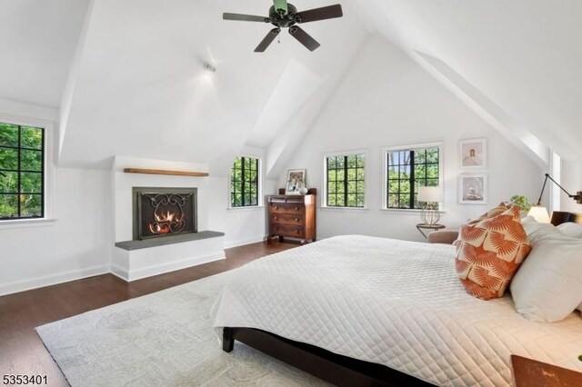 bedroom featuring vaulted ceiling, wood finished floors, baseboards, a warm lit fireplace, and a ceiling fan