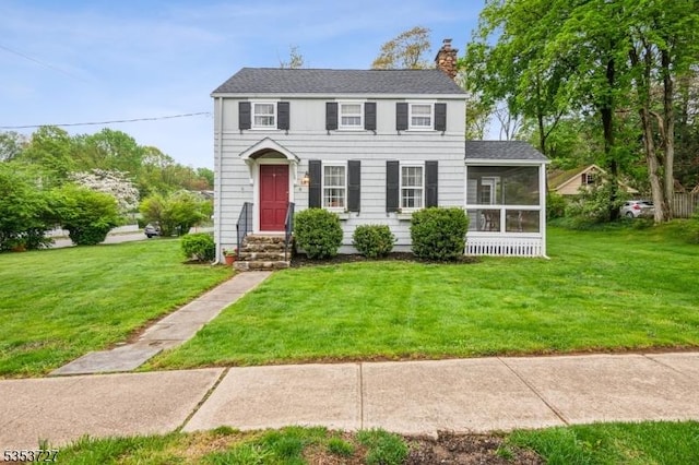 colonial inspired home with a chimney, a front yard, a shingled roof, and a sunroom