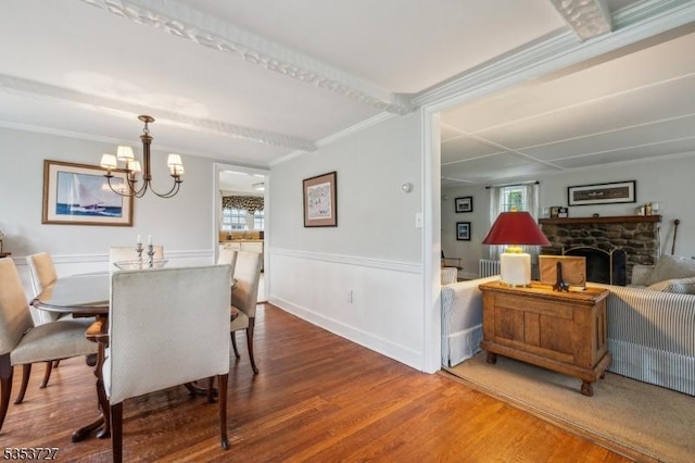 dining space featuring wood finished floors, a stone fireplace, crown molding, a chandelier, and wainscoting