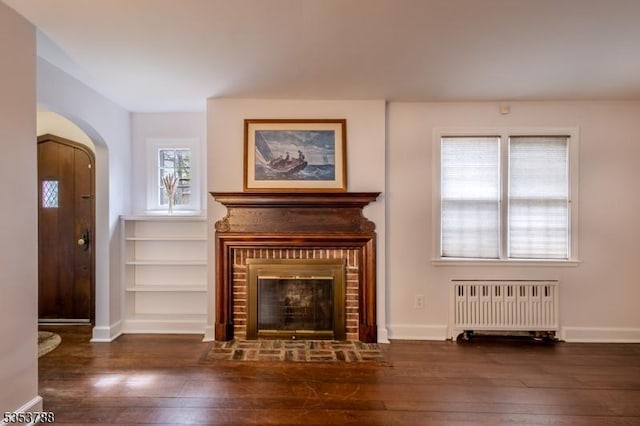 unfurnished living room featuring arched walkways, radiator, a brick fireplace, and hardwood / wood-style flooring
