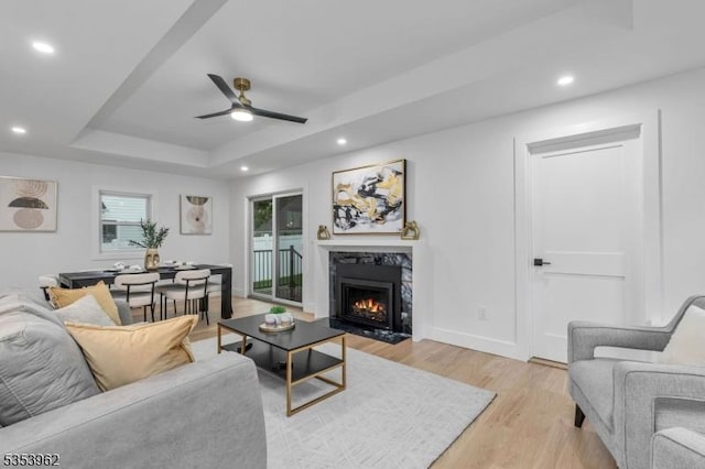 living area with ceiling fan, a raised ceiling, light wood-style floors, a high end fireplace, and recessed lighting