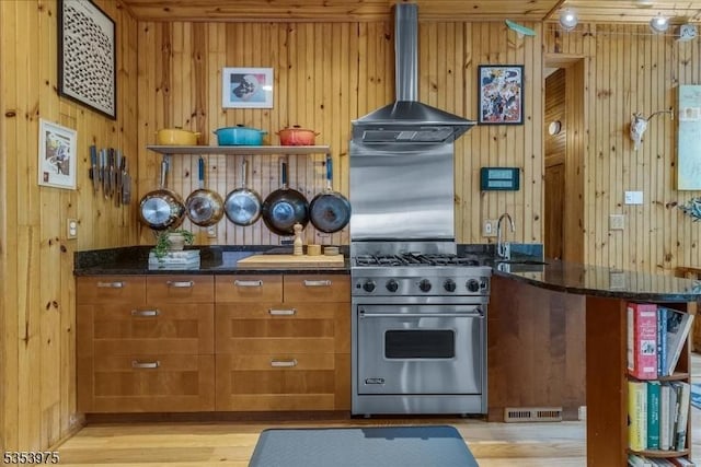kitchen with luxury stove, exhaust hood, dark stone counters, wood walls, and brown cabinetry