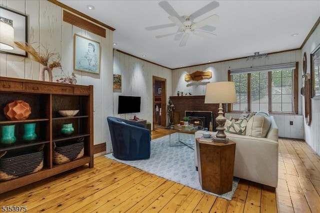 living room featuring wood-type flooring, crown molding, a ceiling fan, wooden walls, and a fireplace