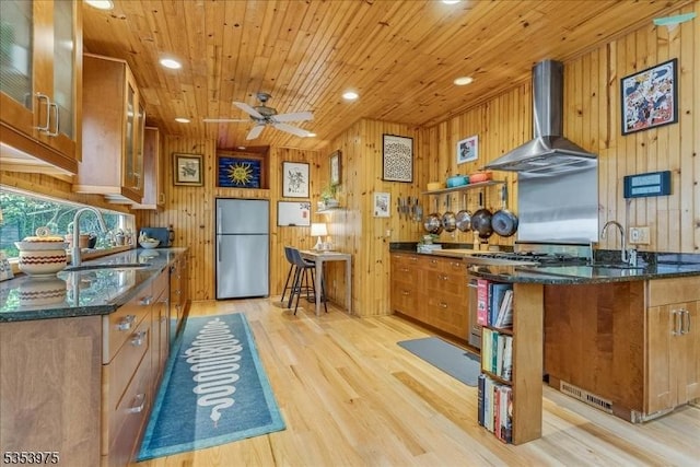 kitchen with stainless steel appliances, wall chimney range hood, a sink, brown cabinets, and wood walls