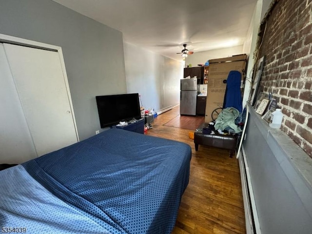 bedroom with ceiling fan, brick wall, freestanding refrigerator, wood finished floors, and a baseboard heating unit